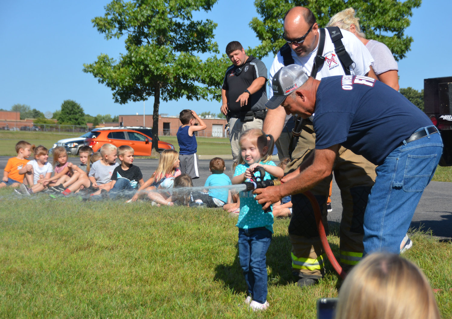 Rylee Shockley, an Owensville Elementary School preschool student, flows water from an Owensville Fire Department  hose Thursday under the supervision of volunteer firemen Assistant Chief Jeff Limberg and Chief Jeff Arnold (in back). Firemen were at the school with other emergency services agencies last week for programs about community leaders.